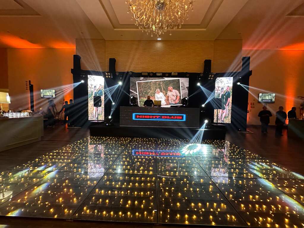 Palco de DJ em uma recepção de casamento, com telas de LED ao fundo, um lustre acima e uma pista de dança iluminada