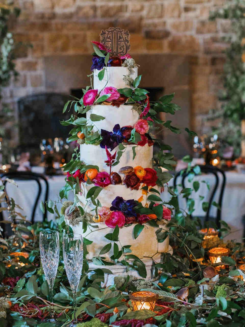 Um bolo de casamento de vários andares decorado com flores coloridas e folhagens verdes, com um topo personalizado. Ao fundo, um cenário rústico de pedra.