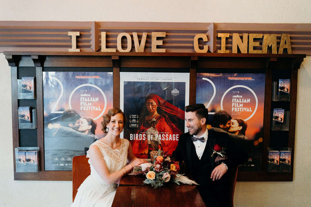 casal de noivos em ensaio dentro de cinema com cartazes de filmes ao fundo