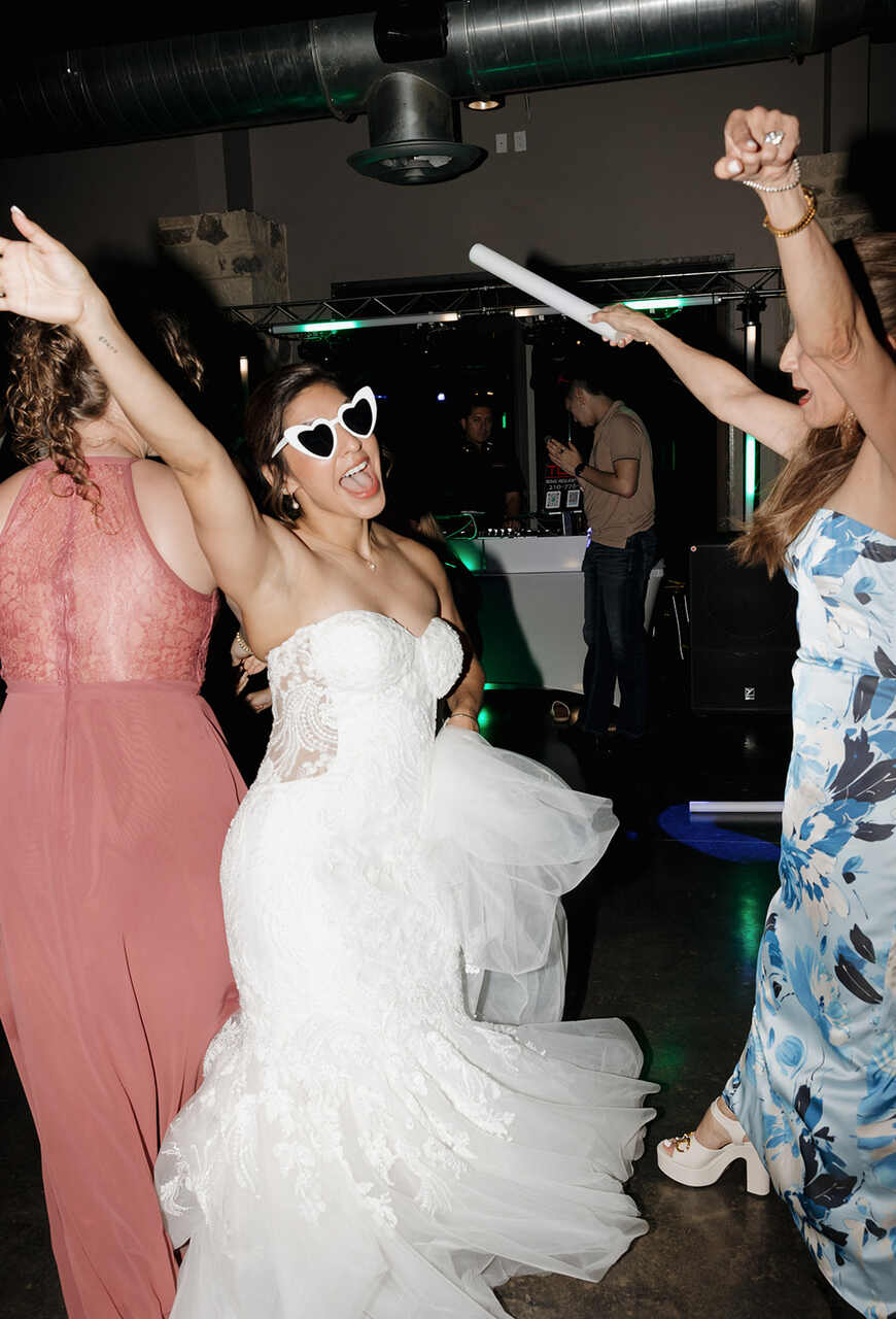 Noiva animada dançando na pista de dança com óculos de coração e vestido de noiva rendado, acompanhada por duas amigas, uma delas com vestido rosa e outra com vestido estampado azul e branco. O ambiente está decorado com luzes verdes, e há um DJ ao fundo.