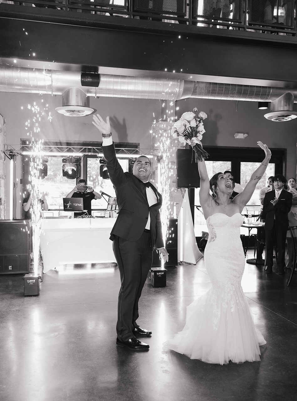 Casal de noivos sorrindo e comemorando com os braços erguidos durante a recepção. A noiva segura um buquê de flores enquanto o noivo também ergue o braço, celebrando a ocasião. Ao fundo, há um DJ e luzes de efeitos especiais.