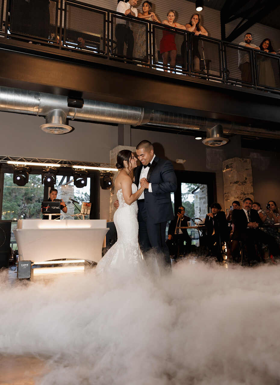 Casal de noivos realizando a primeira dança sobre uma pista coberta de fumaça. O noivo veste um terno escuro e a noiva usa um vestido de noiva detalhado. Ao fundo, o DJ toca em uma mesa iluminada.