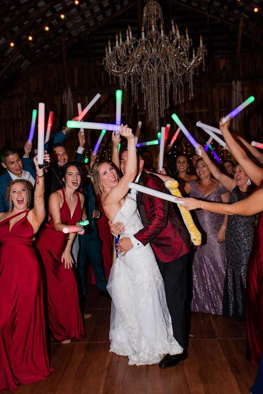 Noivos e convidados animados na pista de dança, segurando bastões de LED coloridos em uma festa de casamento, com um lustre ao fundo