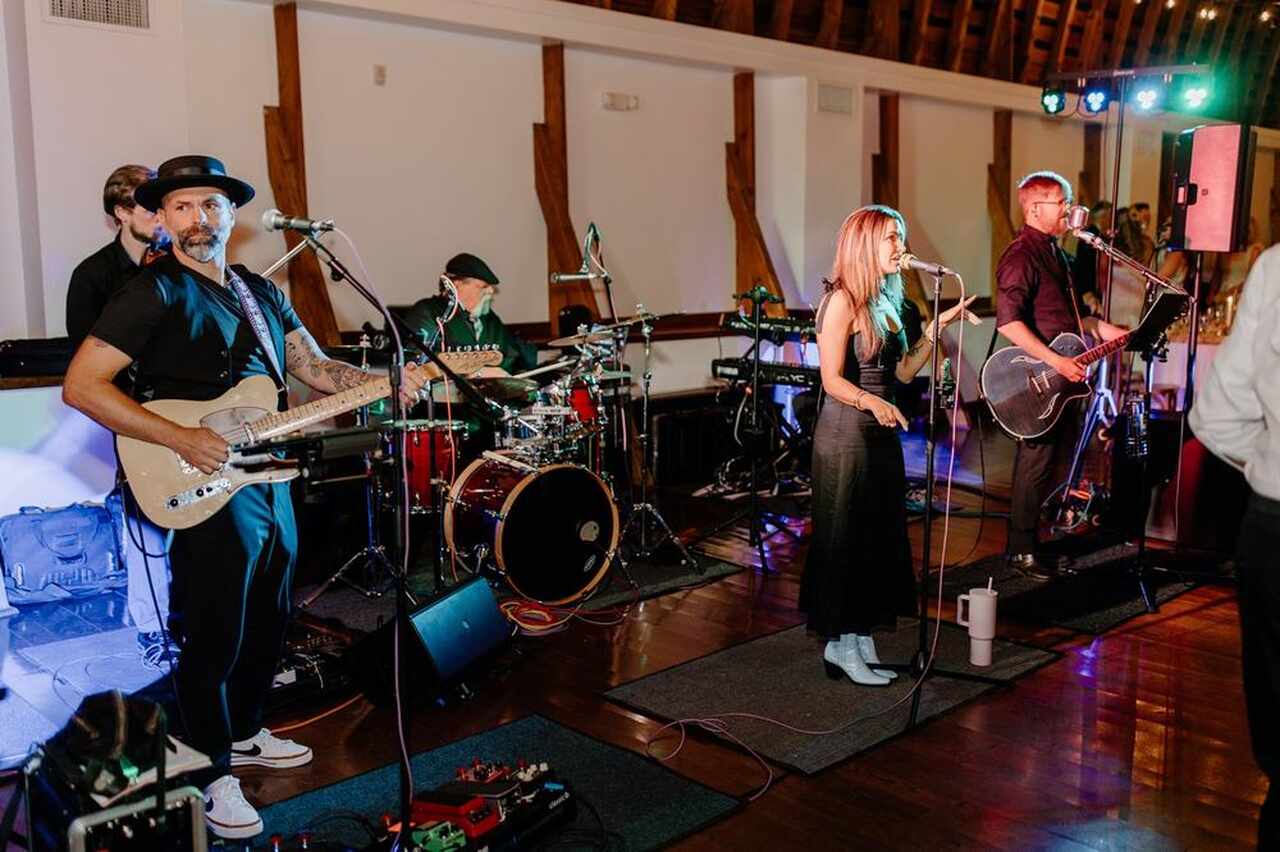 Banda ao vivo tocando durante a festa de casamento. Os músicos, incluindo um guitarrista, baterista, e uma vocalista, estão no palco. A vocalista está vestida de preto e segurando o microfone enquanto canta, com luzes coloridas ao fundo.