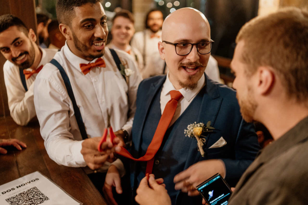 capa post: Noivo com uma gravata laranja, sendo cortada por um dos padrinhos