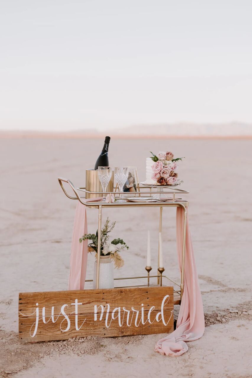 Carrinho dourado decorado com champanhe, flores e uma placa "Just Married", em um cenário desértico minimalista.