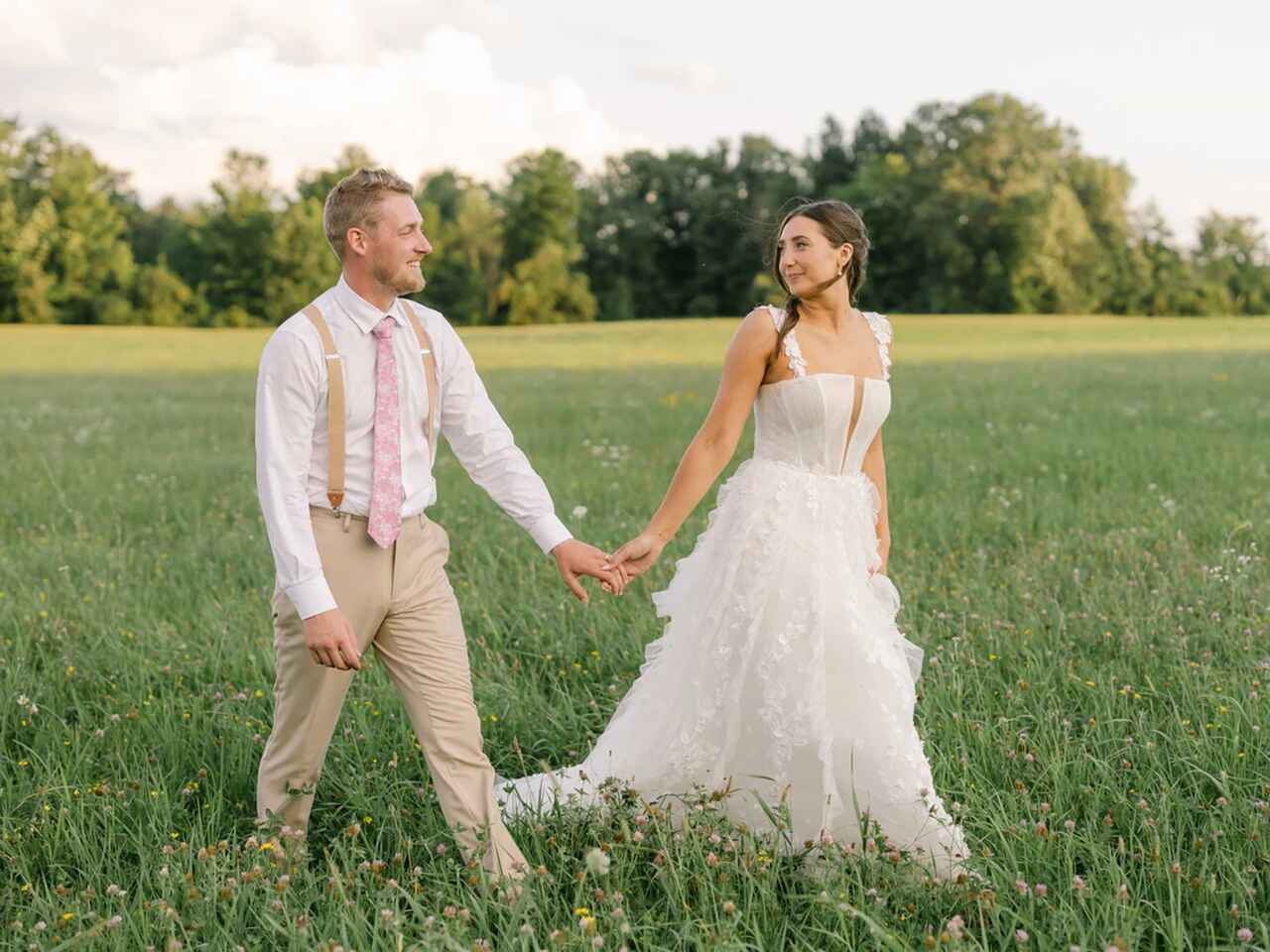 Casal de noivos caminhando de mãos dadas em um campo verde, com a noiva em um vestido branco de alças e o noivo em suspensórios bege e gravata rosa.