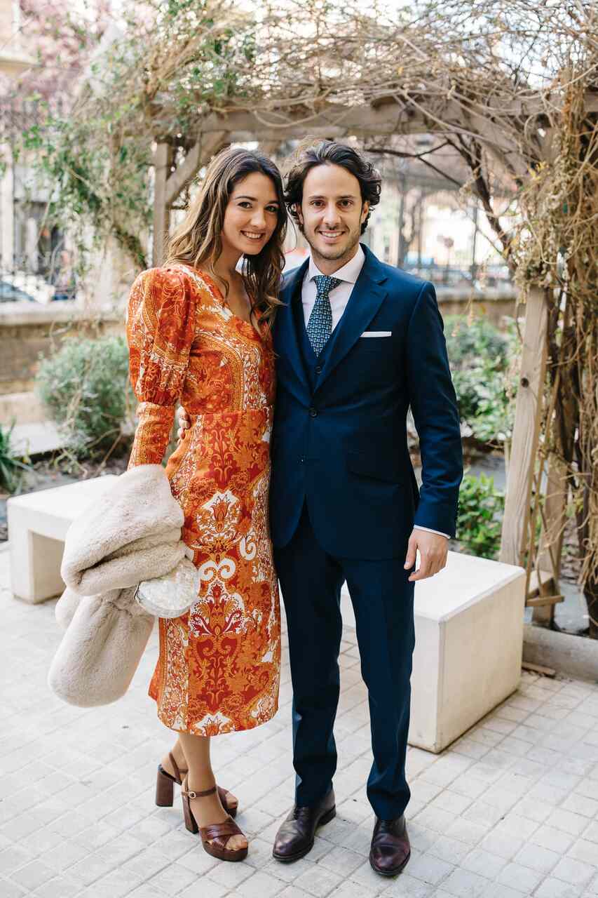 Casal em casamento ao ar livre; ele de terno azul e gravata, ela em vestido estampado vermelho e dourado com detalhes sofisticados.