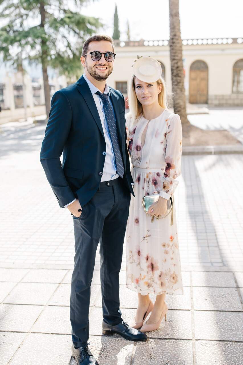 Casal elegante posando em local ensolarado; ele com terno azul e gravata, ela em vestido floral e chapéu discreto.