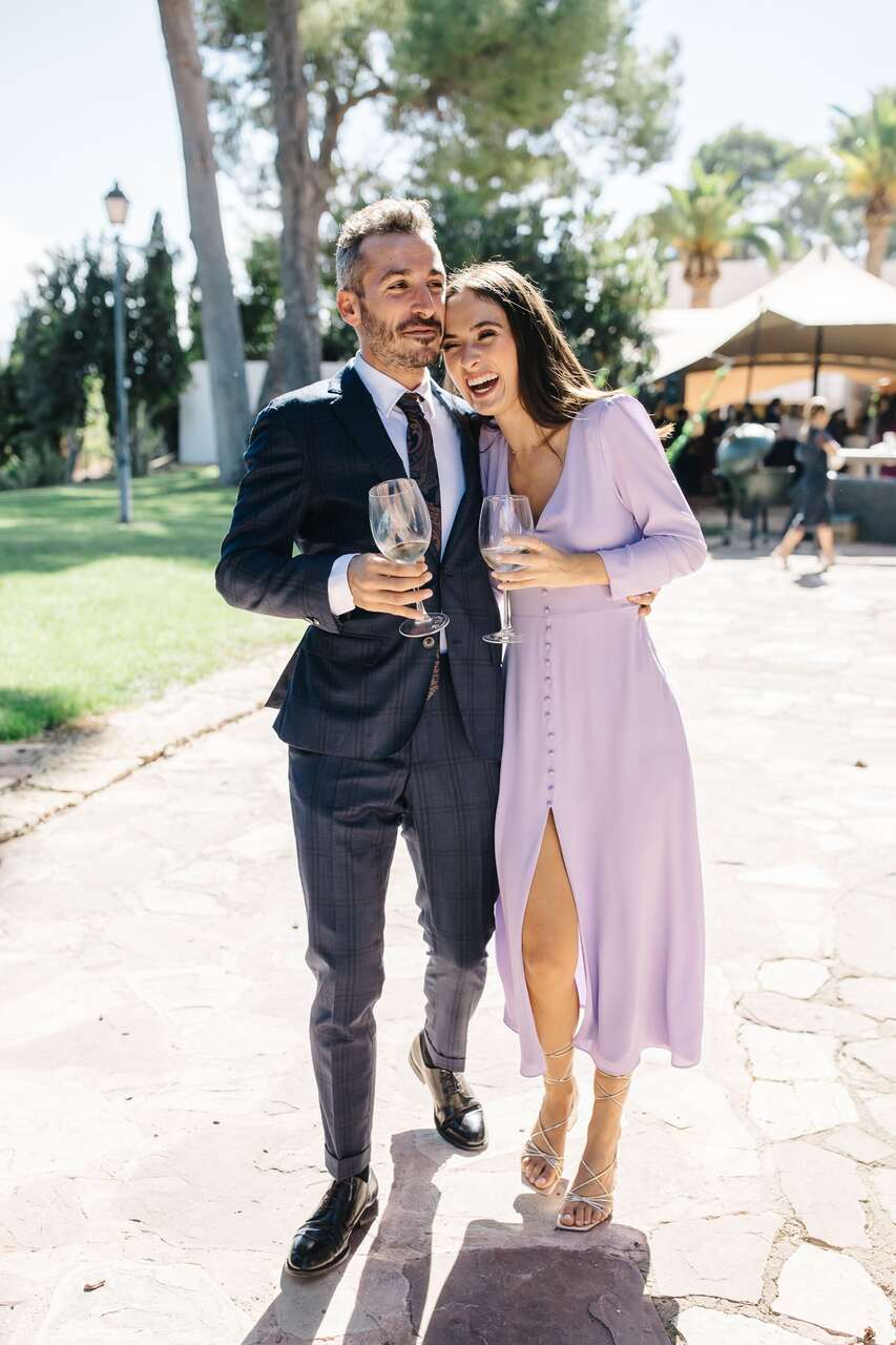 Casal sorrindo em um casamento ao ar livre; ele em terno azul escuro, ela em vestido lilás com taças de vinho na mão.