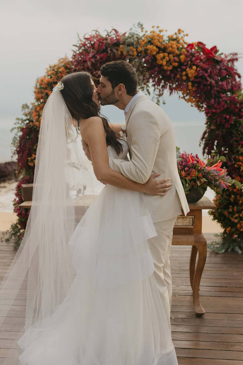 Casal se beijando em frente a um arco de flores coloridas em um casamento ao ar livre.