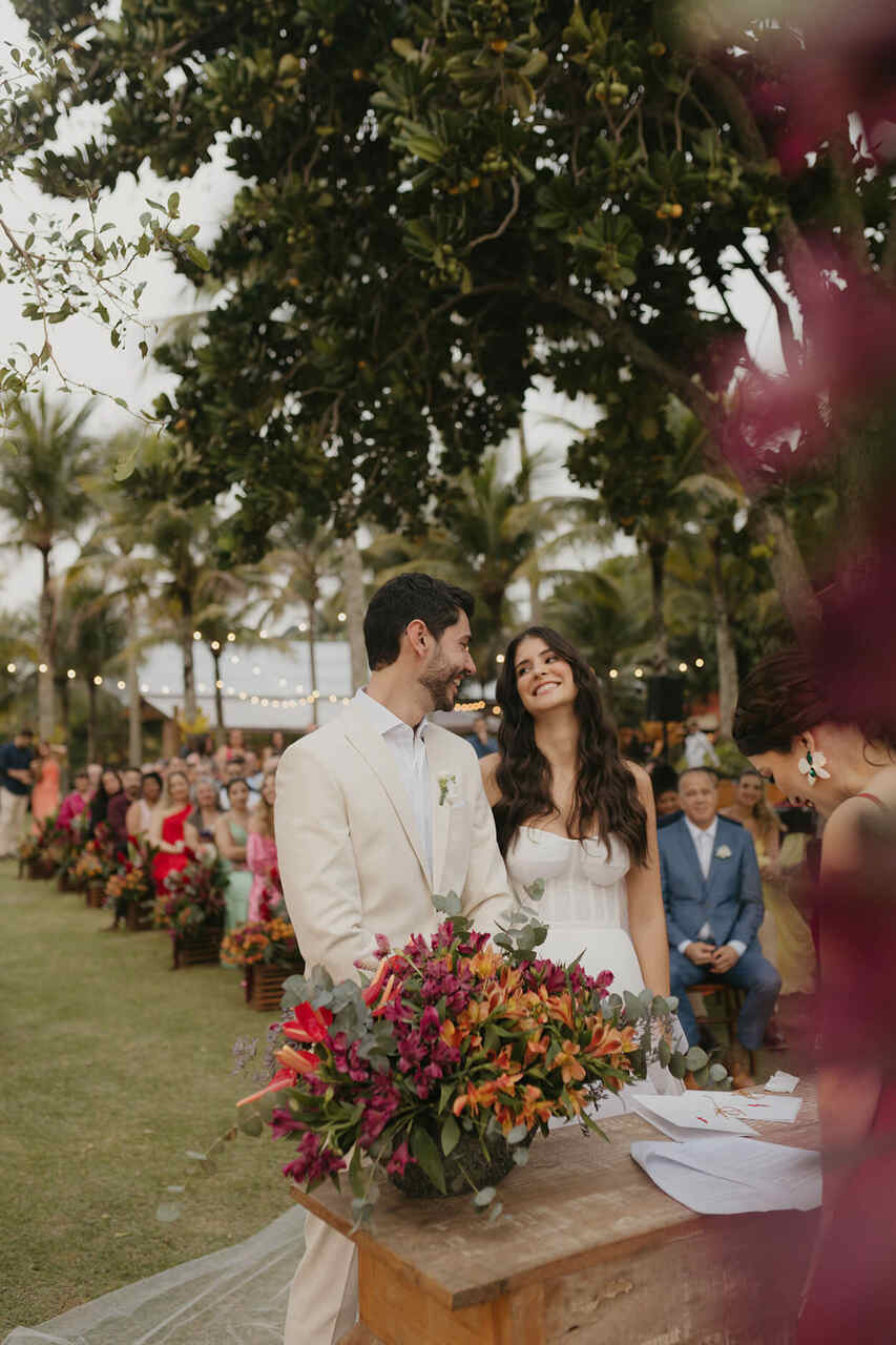 Casal sorrindo durante a cerimônia ao ar livre, rodeado por arranjos florais vibrantes e convidados sentados, em um casamento tropical intimista