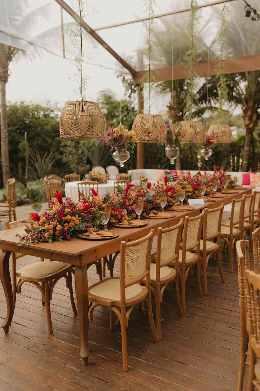 Mesa de casamento tropical com decoração em tons quentes, arranjos florais vibrantes e luminárias de bambu suspensas em um ambiente ao ar livre com tema rústico e tropical.