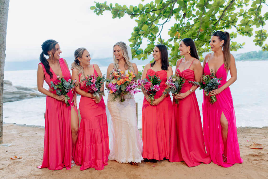 Noiva rodeada por suas madrinhas vestidas em tons de rosa vibrante, todas segurando buquês coloridos, em uma praia