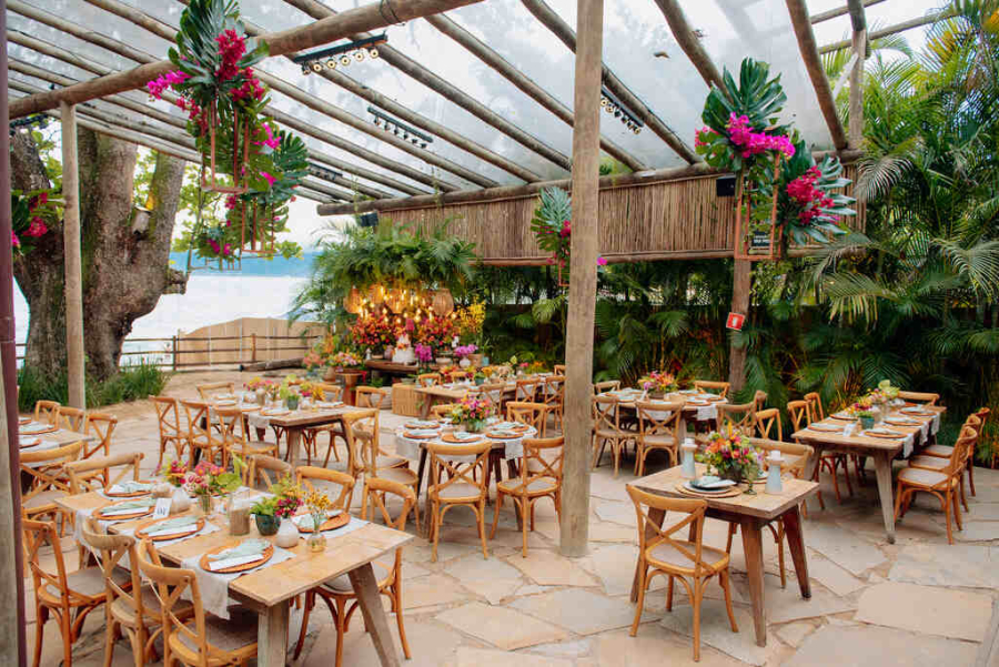 Espaço ao ar livre com mesas de madeira decoradas com arranjos florais tropicais e teto de vidro com plantas pendentes