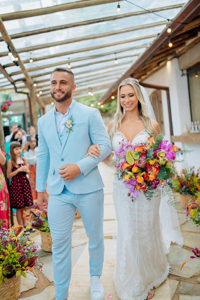 Casal caminhando sorridente, com noivo de terno azul claro e noiva com buquê colorido, em uma cerimônia ao ar livre.