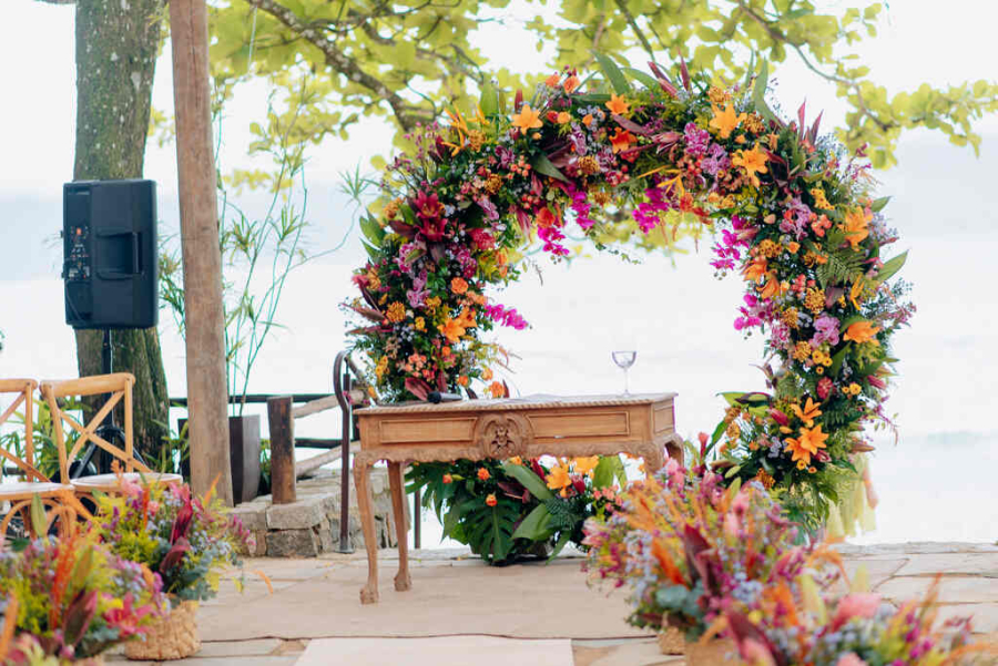 Arco floral vibrante em tons tropicais, emoldurando uma mesa rústica, com vista para o mar ao fundo