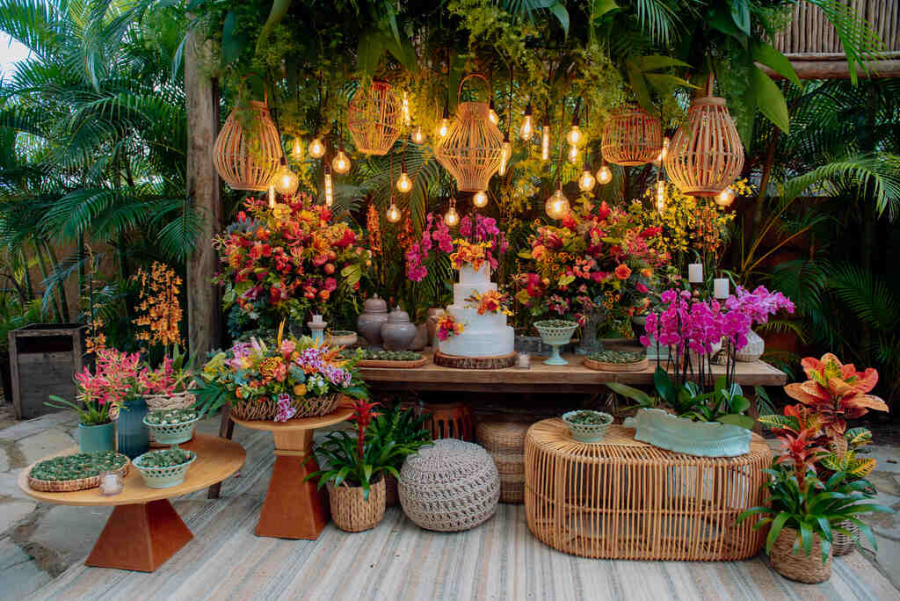 Mesa de bolo decorada com flores tropicais, luminárias de palha suspensas e elementos rústicos em um ambiente ao ar livre.