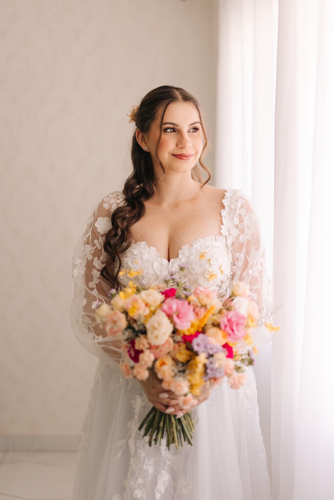 Uma noiva em um vestido branco com detalhes florais segura um buquê colorido de flores em tons pastel, enquanto olha serenamente pela janela iluminada.