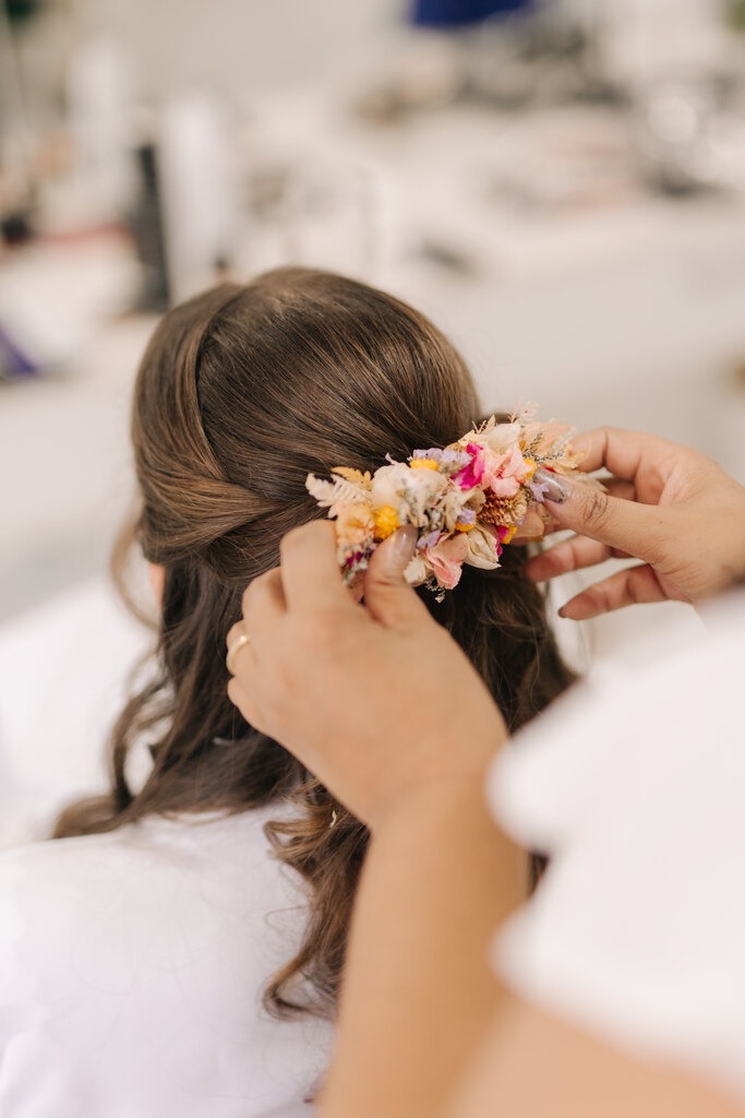 Detalhe de uma noiva recebendo um acessório floral colorido em seu penteado, enquanto se prepara para o casamento.