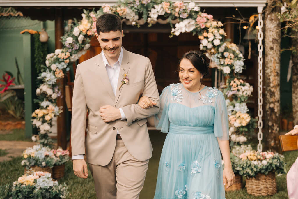  Homem de terno bege acompanhando uma mulher com vestido azul claro bordado, caminhando sob um arco floral.