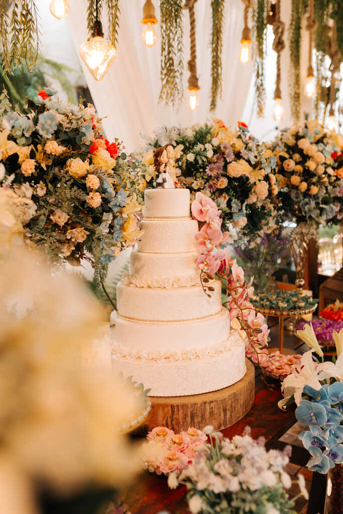 Bolo de casamento de cinco andares com acabamento perolado, decorado com flores em tons de rosa e branco, posicionado em uma mesa rústica de madeira cercada por exuberantes arranjos florais em um ambiente iluminado com lâmpadas pendentes.