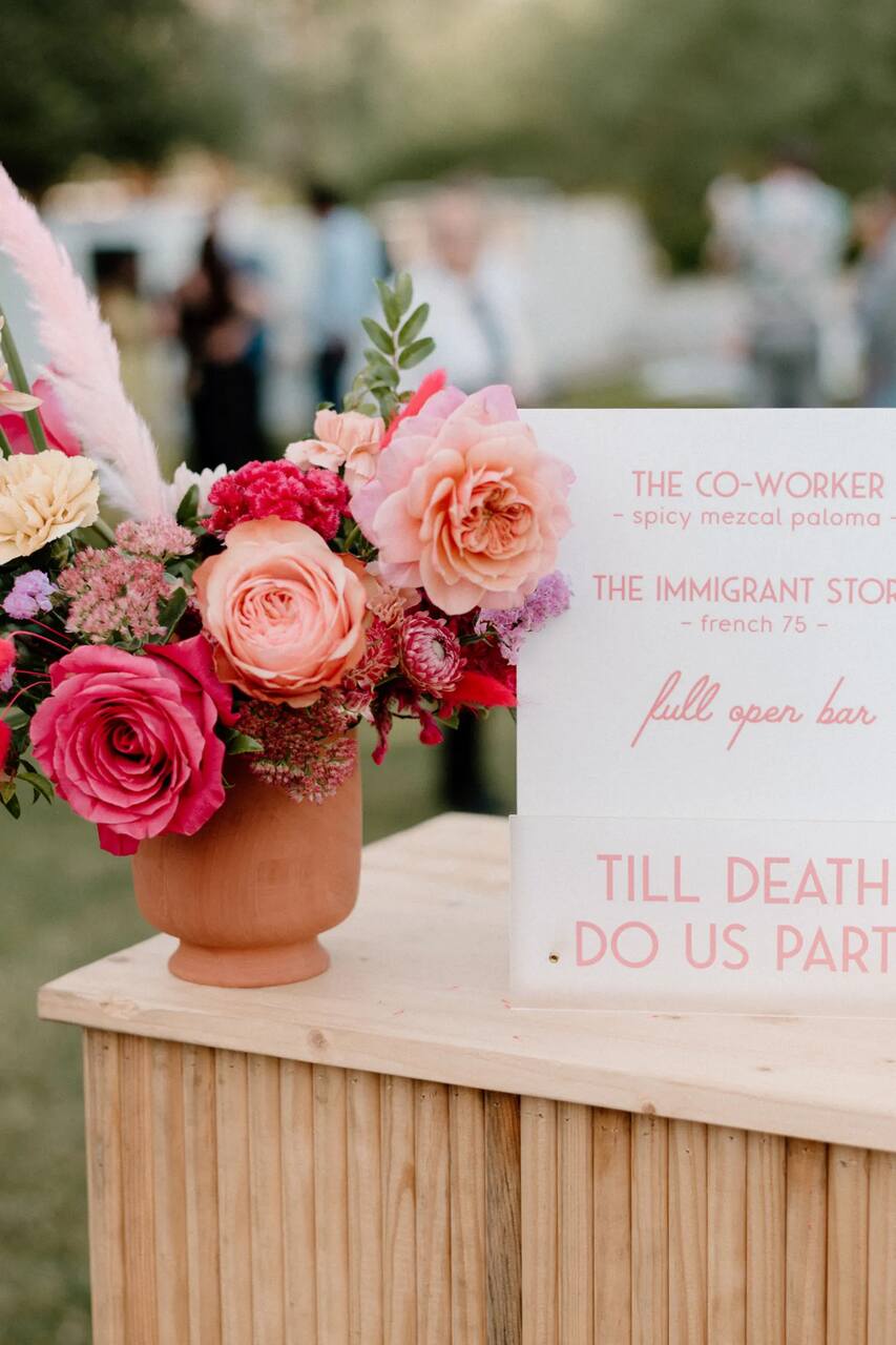 Arranjo floral com rosas vibrantes ao lado de uma placa de menu do bar, compondo uma decoração tropical para um casamento ao ar livre