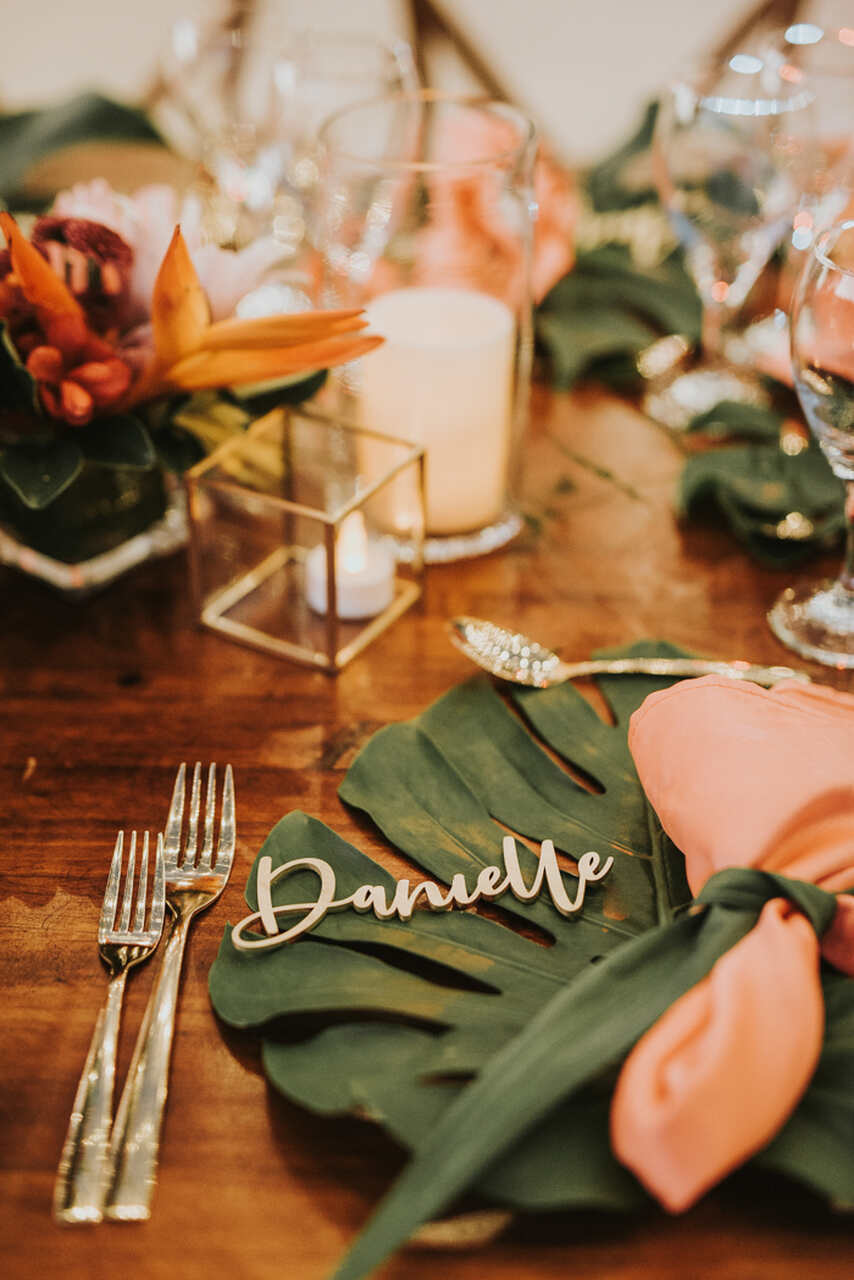 Lugar à mesa com folha de costela-de-adão, guardanapo rosa e nome personalizado, decorado com arranjos tropicais e velas