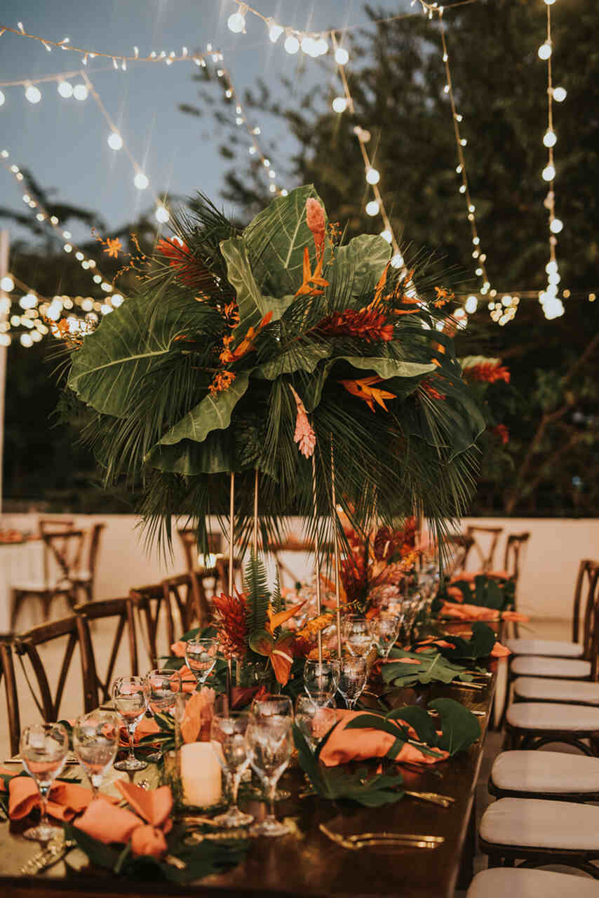 Mesa de jantar ao ar livre com arranjos altos de folhas tropicais e flores vibrantes, iluminada por luzes de cordão no entardecer