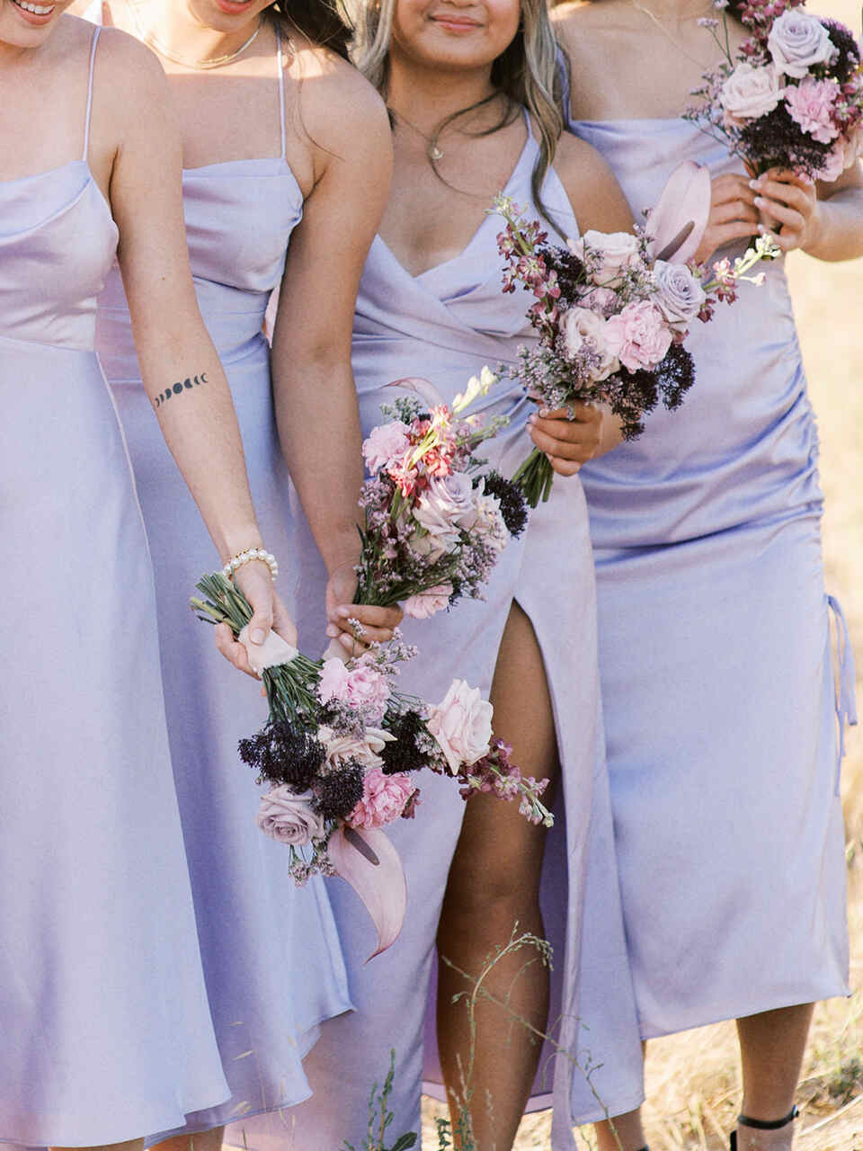 Damas de honra vestindo vestidos lilás claros segurando delicados buquês em tons de rosa e lavanda, destacando a harmonia das cores no cenário ensolarado.