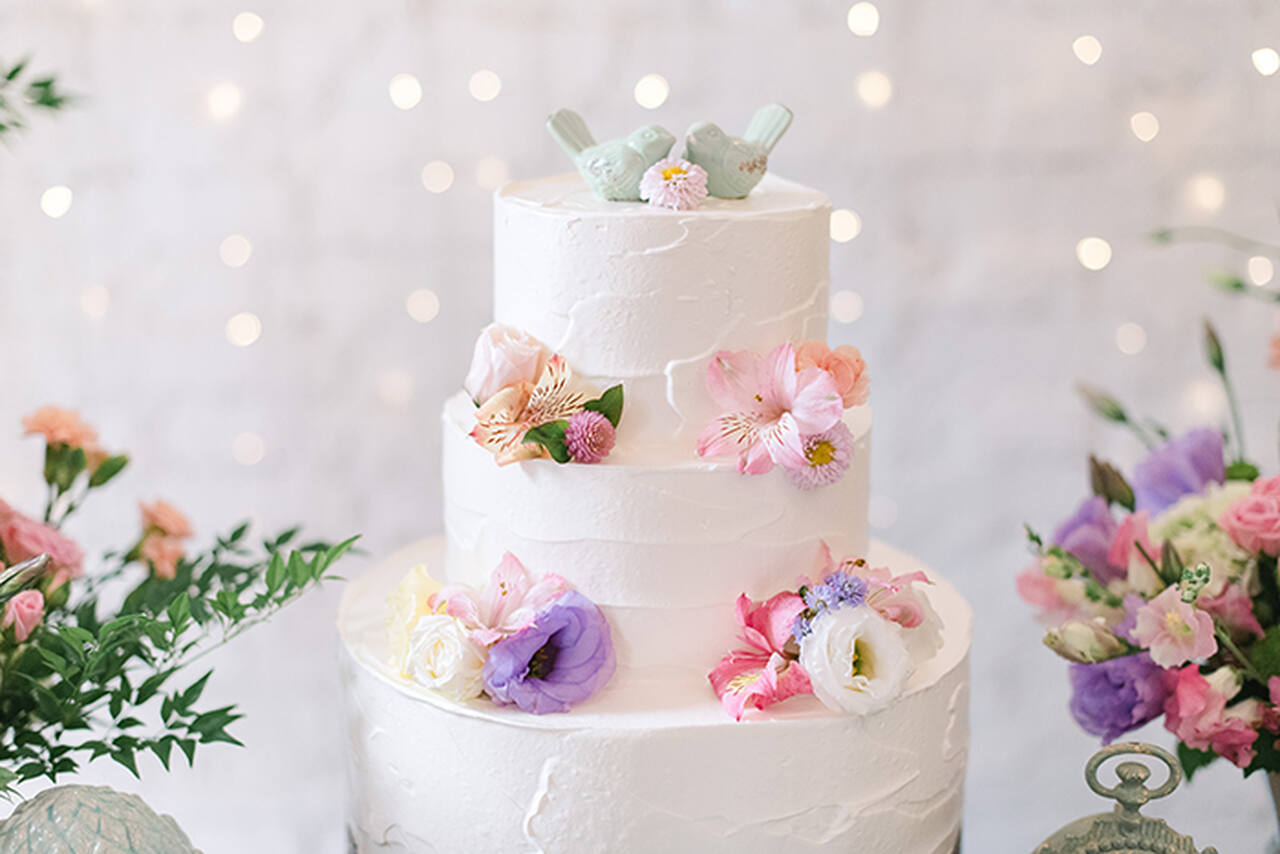 olo de casamento branco de três andares, decorado com flores delicadas em tons de rosa, lilás e branco. No topo, dois passarinhos decorativos verdes complementam o design romântico. O fundo é iluminado por luzes suaves 