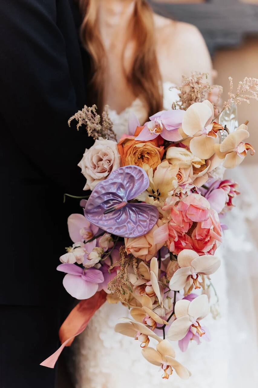 Um close do buquê mostra a riqueza de detalhes com flores como antúrios lilás, rosas em tons quentes e em tons pastel, delicados toques de orquídeas