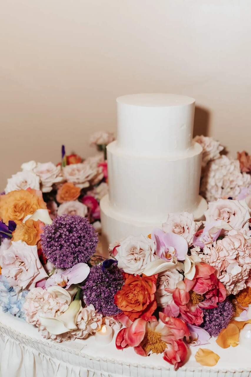 Bolo de casamento minimalista de três andares, com acabamento liso e sem decoração adicional, cercado por um exuberante arranjo de flores vibrantes em tons de laranja, roxo, rosa e creme