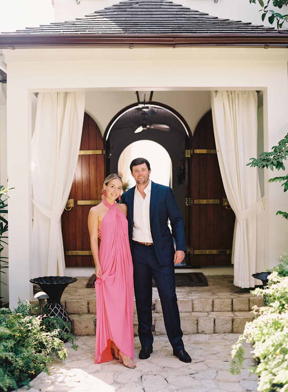 asal elegante posando em frente a um portão decorado; homem com terno azul marinho e mulher em vestido longo rosa claro