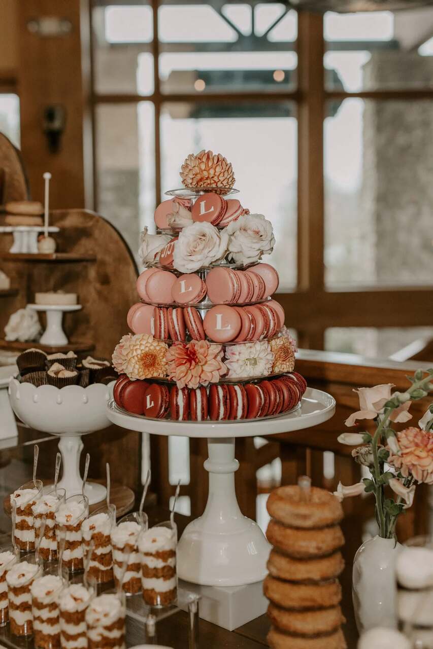 Torre de macarons rosa organizados em camadas, decorados com flores frescas e detalhes personalizados com a letra "L", compondo uma elegante mesa de doces com outras sobremesas ao fundo.