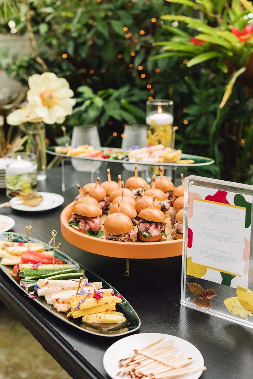 Mesa de buffet com mini hambúrgueres e petiscos variados em um ambiente decorado com plantas.