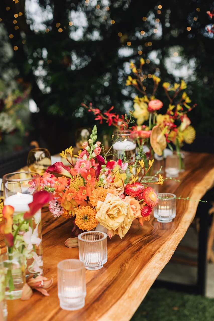 Mesa de madeira rústica decorada com flores vibrantes em tons quentes e velas em recipientes de vidro