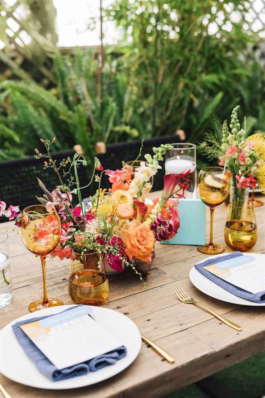 Mesa ao ar livre decorada com arranjos florais coloridos, taças âmbar e detalhes elegantes em um ambiente natural