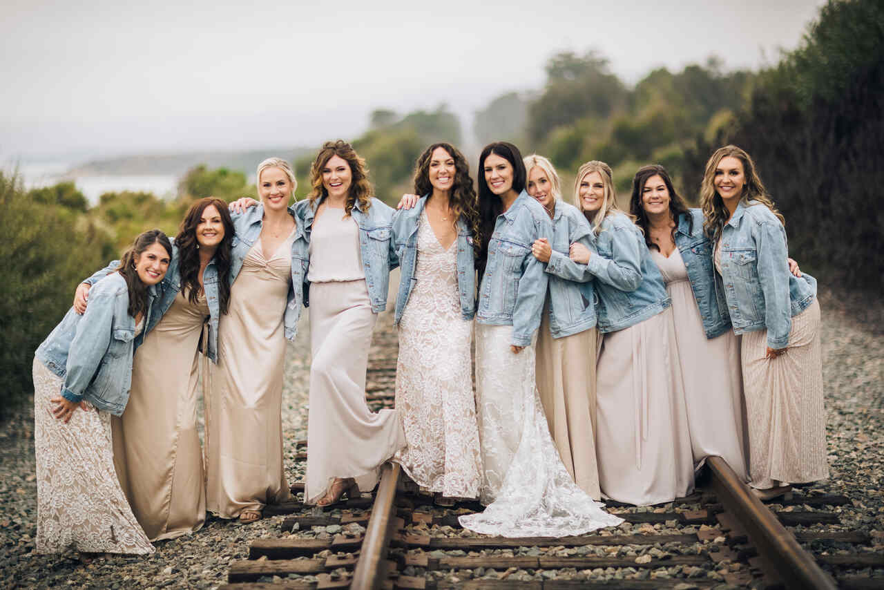 Noiva e suas madrinhas em vestidos neutros, combinados com jaquetas jeans, posando descontraídas sobre trilhos de trem.