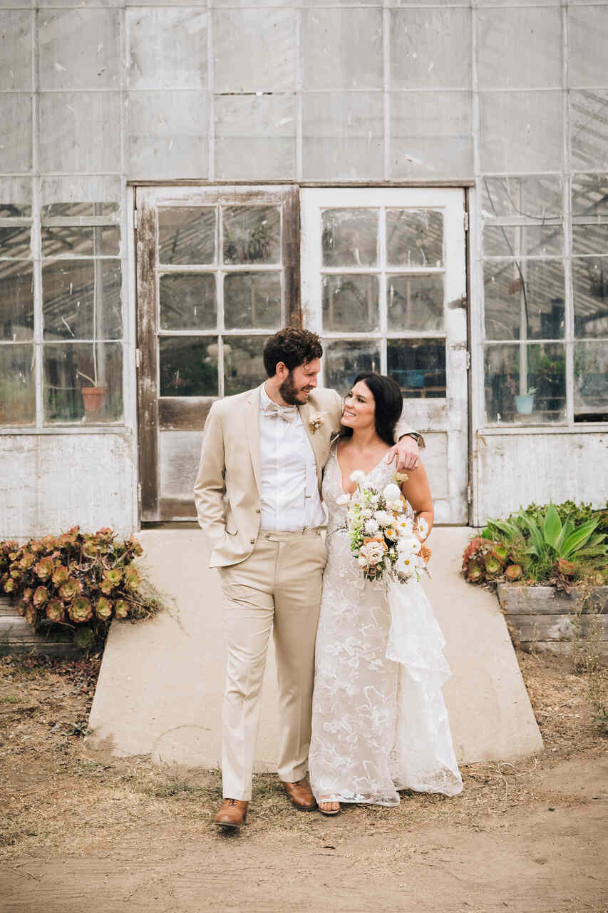 Casal sorridente em frente a uma estufa rústica, a noiva em um vestido rendado e o noivo em um terno bege com gravata borboleta.