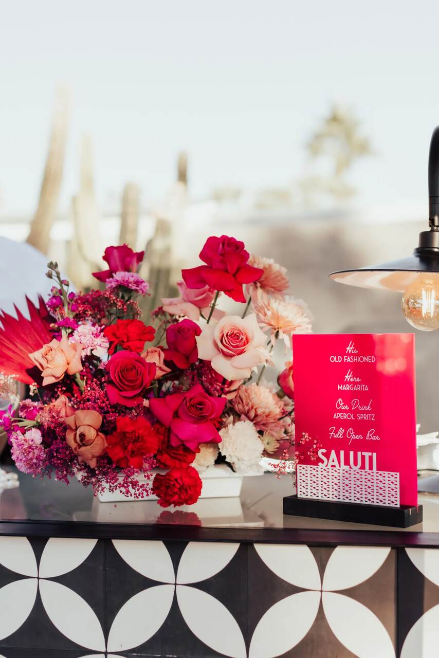 Balcão de drinks com arranjo de flores vermelhas e rosa, acompanhado por um cardápio de coquetéis em destaque