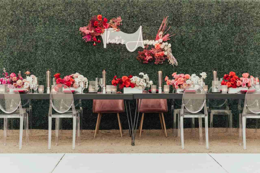 Mesa de jantar sofisticada decorada com arranjos florais em tons de vermelho e rosa, com fundo de parede verde e letreiro 'That's Amore