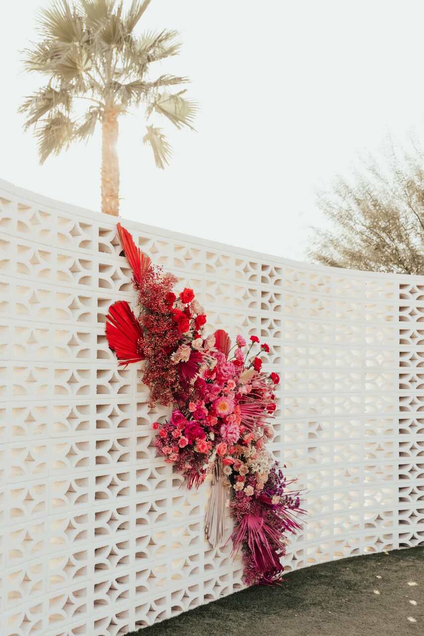 Mural branco com um arranjo vertical de flores vibrantes em tons de rosa e vermelho, destacando-se sob uma palmeira