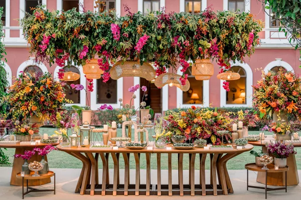 Mesa decorada com flores coloridas e iluminação suspensa em um evento ao ar livre.