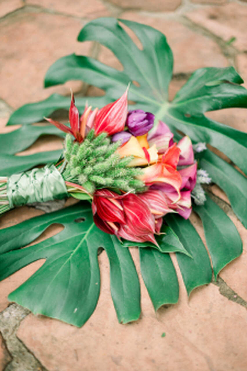 Buquê tropical com flores exóticas em tons quentes, disposto sobre folhas grandes de costela-de-adão.