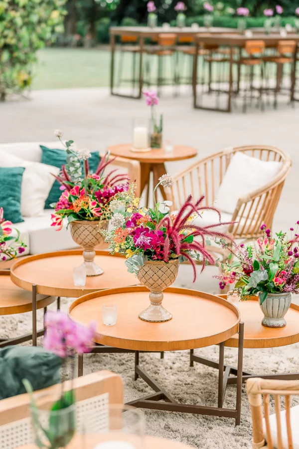 Lounge vibrante com mesas redondas de madeira, arranjos florais coloridos e cadeiras em estilo rústico