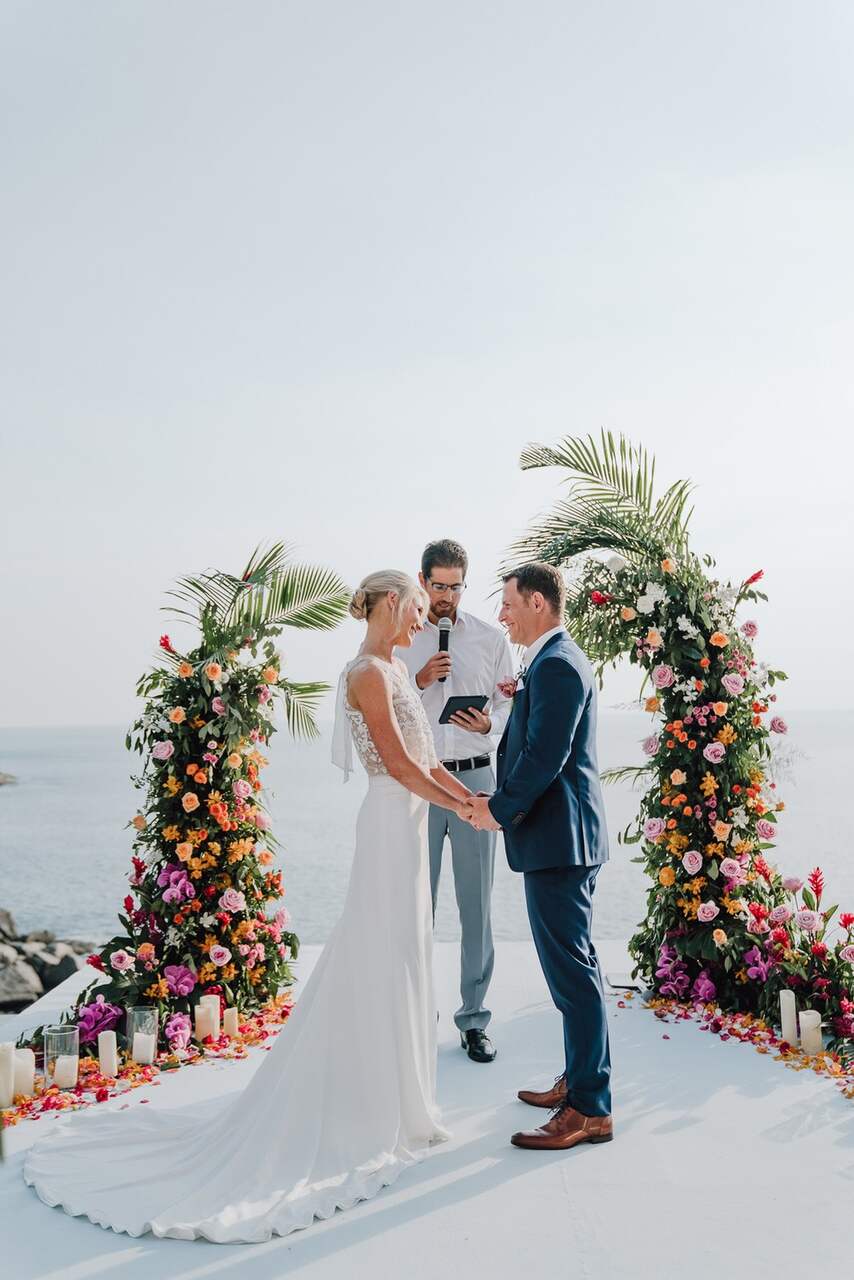 Casal de noivos se olhando carinhosamente durante a cerimônia de casamento ao ar livre, com o mar e um arco decorado com flores vibrantes ao fundo.