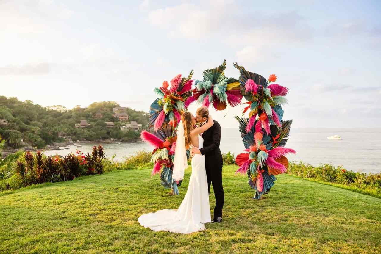 Casal se beijando em um casamento tropical ao ar livre, com um arco decorado com folhas e flores coloridas, em um gramado com vista para o mar