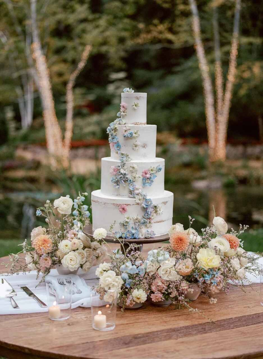 Bolo de casamento alto com cinco camadas, adornado com flores coloridas em tons pastel, cercado por arranjos florais sobre uma mesa rústica ao ar livre.