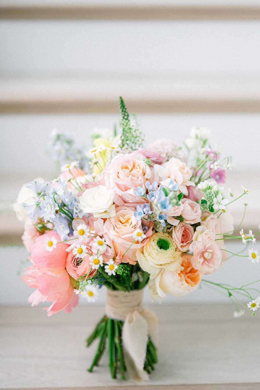 Um close de um delicado buquê de casamento com flores em tons suaves de rosa, pêssego e creme, amarrado com uma elegante fita